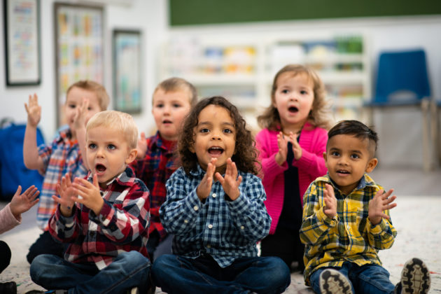 A group of multi-ethnic preschool students sit on a classroom rug. They are singing, doing actions, clapping and having fun during a sing-along time. Each is wearing casual clothing in bright colors.