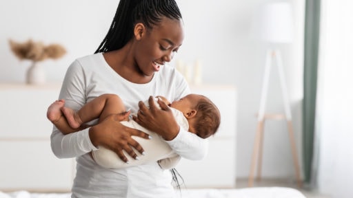 Portrait of smiling African American woman holding her adorable small black upset baby on hands, singing lullaby to sleep, comforting infant kid standing in bedroom at home