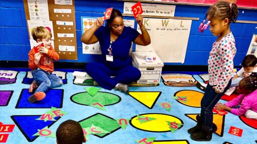 Ms. Williams is sitting on a floor rug with several children. The rug is a light blue with different colored shapes. There are circles, triangles, squares, and diamond shapes on the rug.