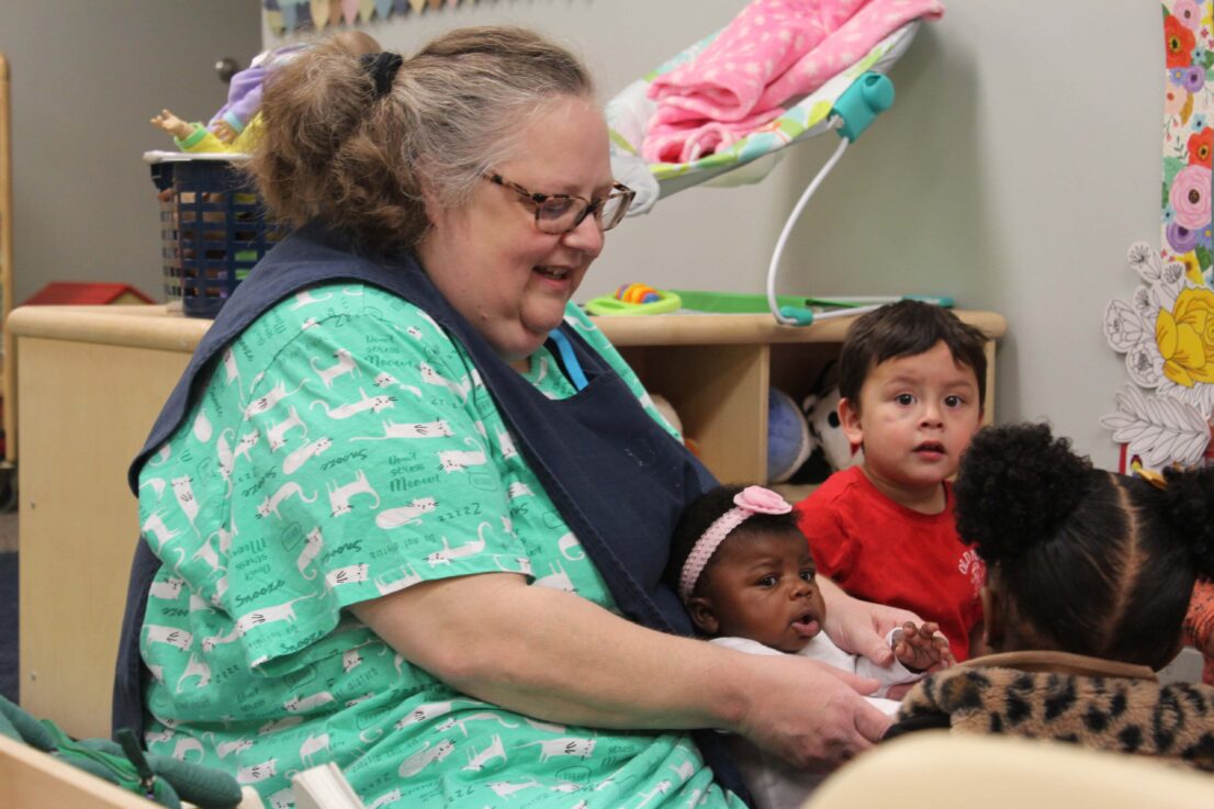 A caregiver wearing a green shirt with cat designs is seated in a daycare environment, holding a toddler on their lap. Two other young children sit close by, engaged in the moment. Behind them, a shelf filled with toys and books and a vibrant wall display add a playful atmosphere to the room.