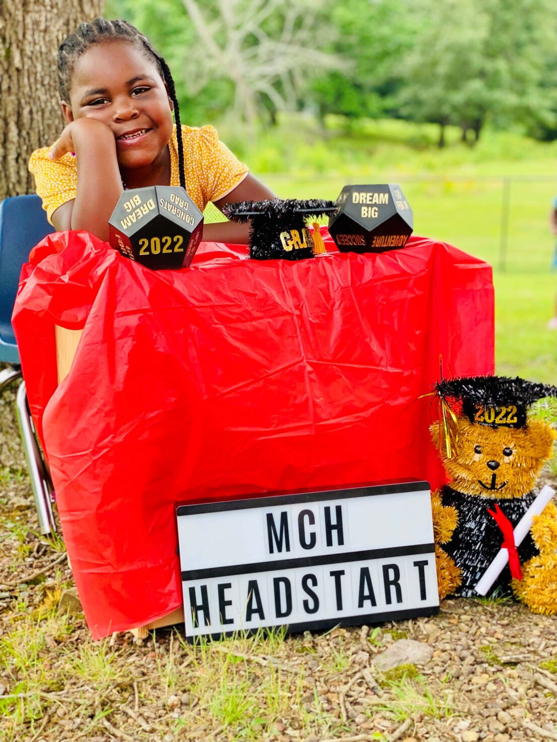 Girl smiling and sitting behind a desk with decorations for graduation and a sign at the bottom of the table saying MCH Head Start