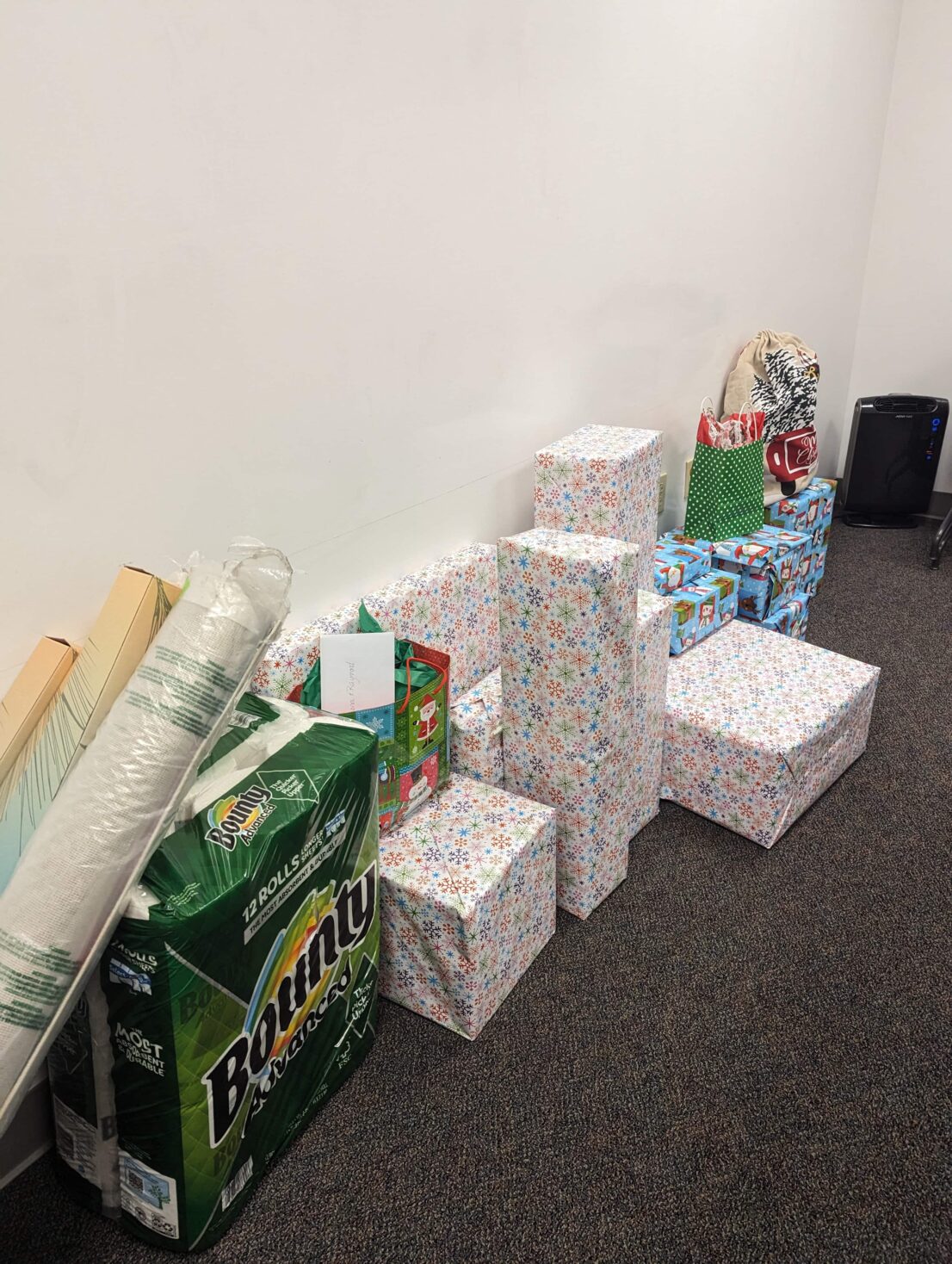 Two long tall boxes and a rolled up rug in plastic lean on a 12 pack of paper towels. Next to this there are about 15 gifts wrapped in white snowflake and blue santa wrapping paper. On top of two green gift bags. One has santa and snow men on it and the other has whilte polka dots. At the far end there is a brown fabric gift bag. Everything is lined up against the wall.