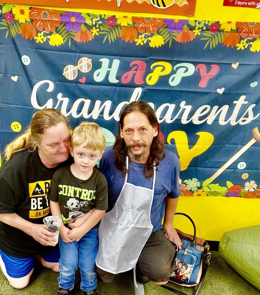 grandparents pose with grandson in front of grandparent's day sign