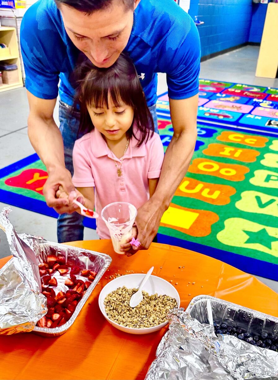 man stands with child as they both hold a spoon and scoop as strawberry into a cup