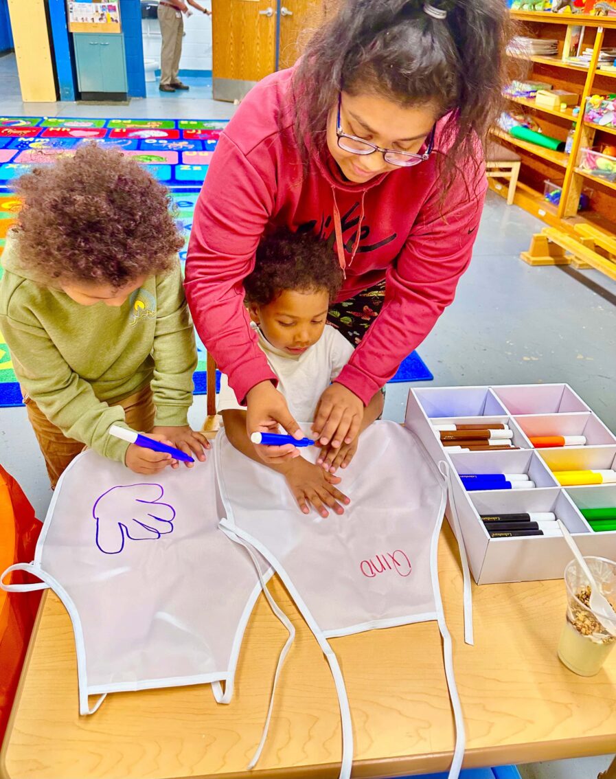 woman traces child's hand while other child draws on apron