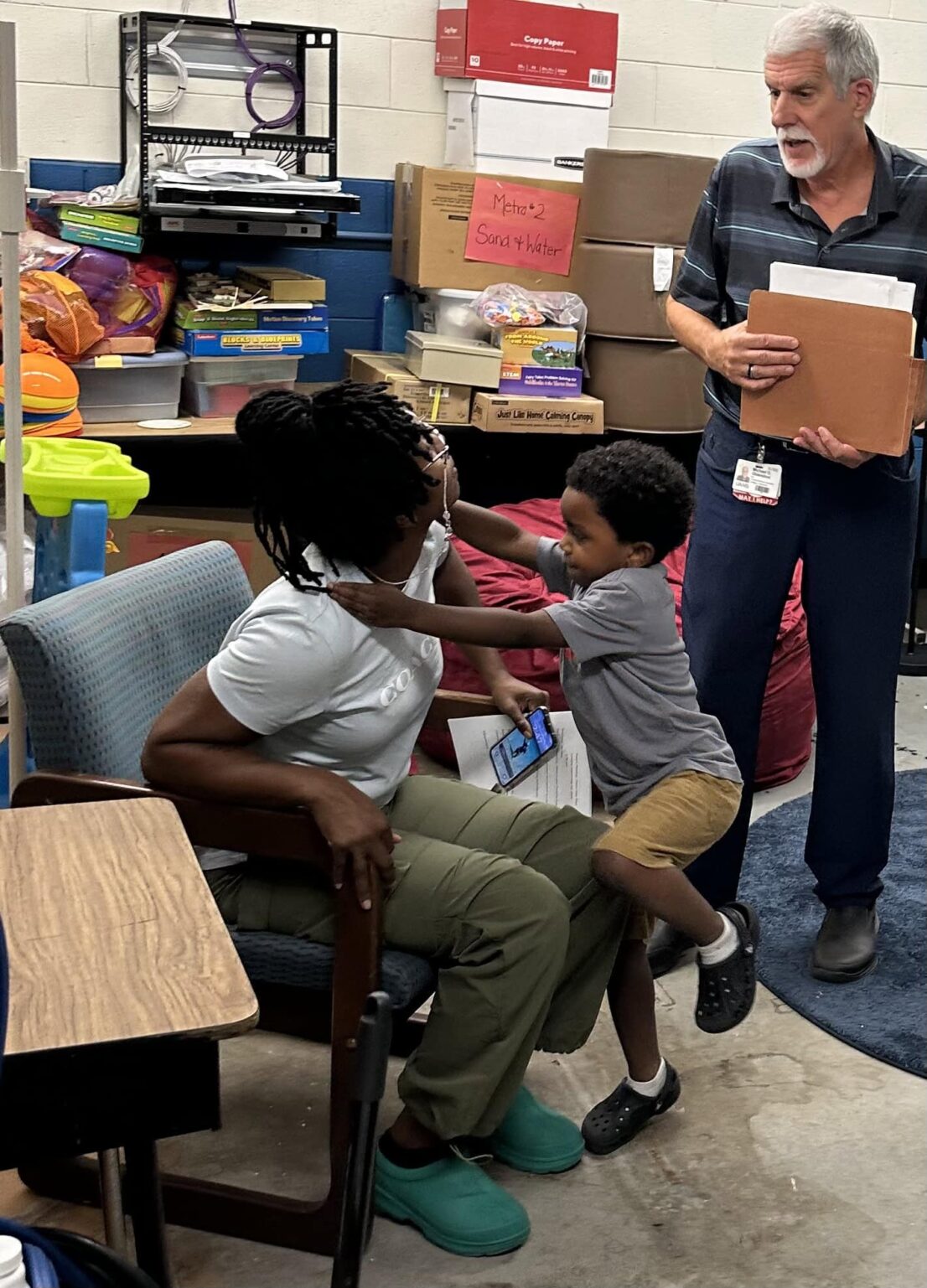 parent sitting in a chair with child climbing onto her lap while a staff person is standing and talking to her