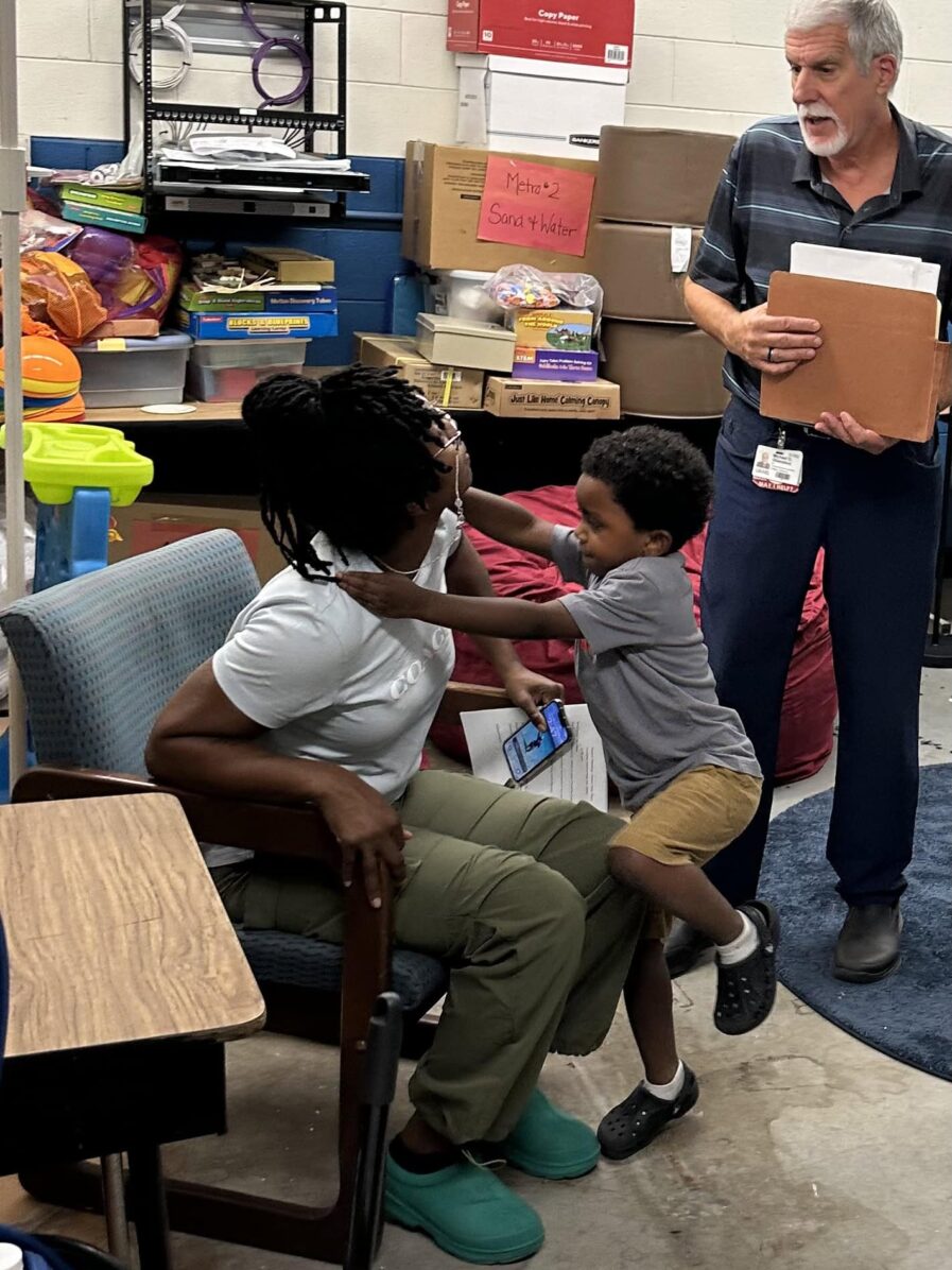 parent sitting in a chair with child climbing onto her lap while a staff person is standing and talking to her