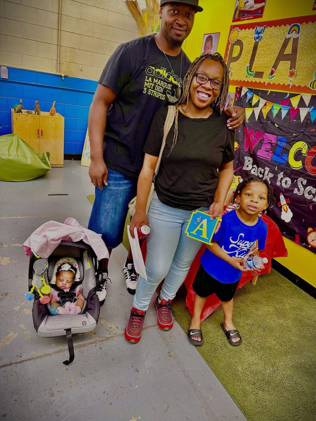 A mother and father stand with a child and a baby is in a car seat that is on the floor next to them.