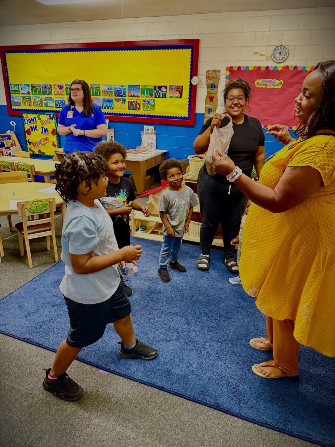 An adult is in the foreground engaging a group of young children while 2 other adults look on in the background