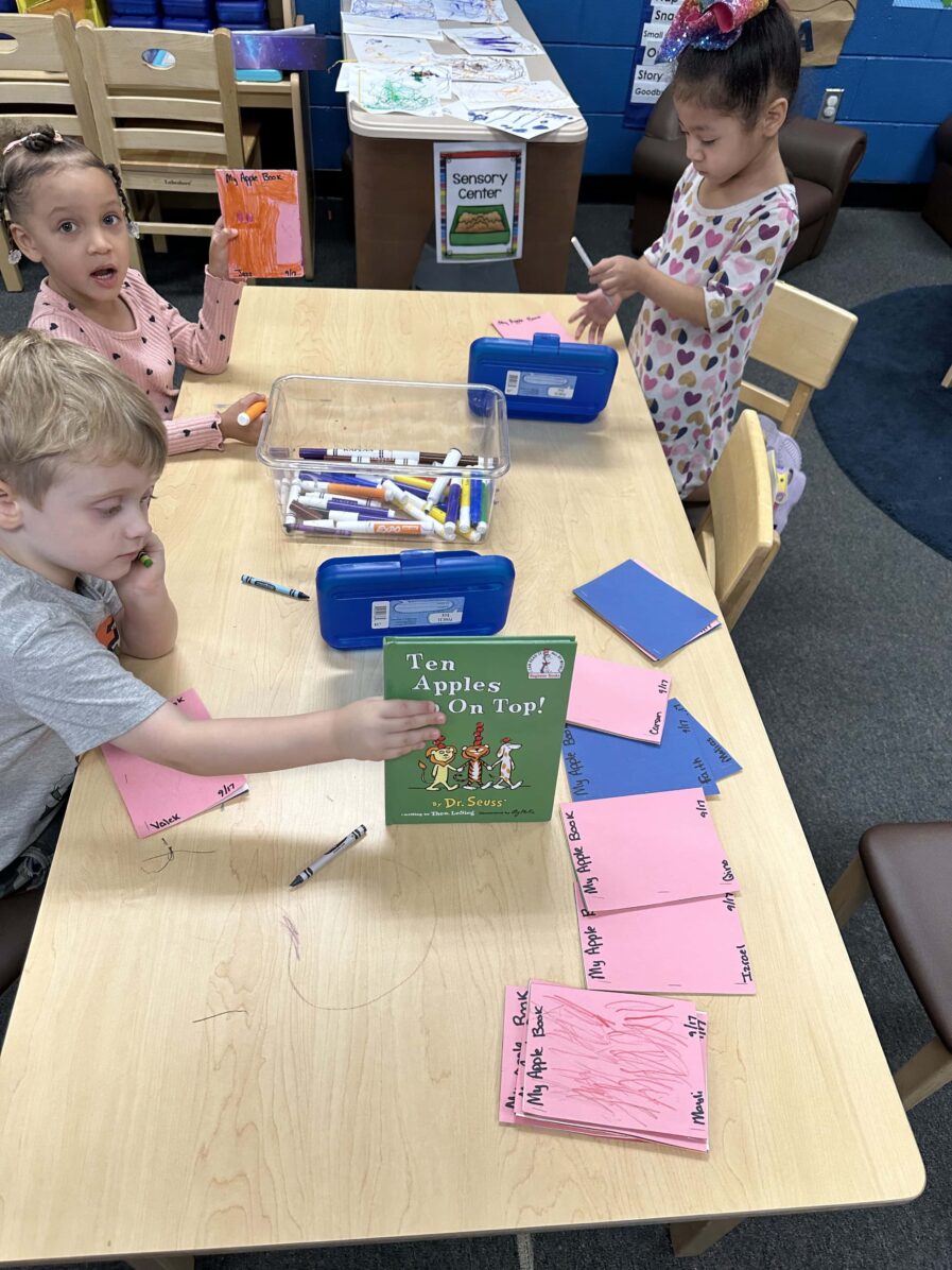 3 children sit around a long table, one child is holding up a book. On the table are child-made books about apples.