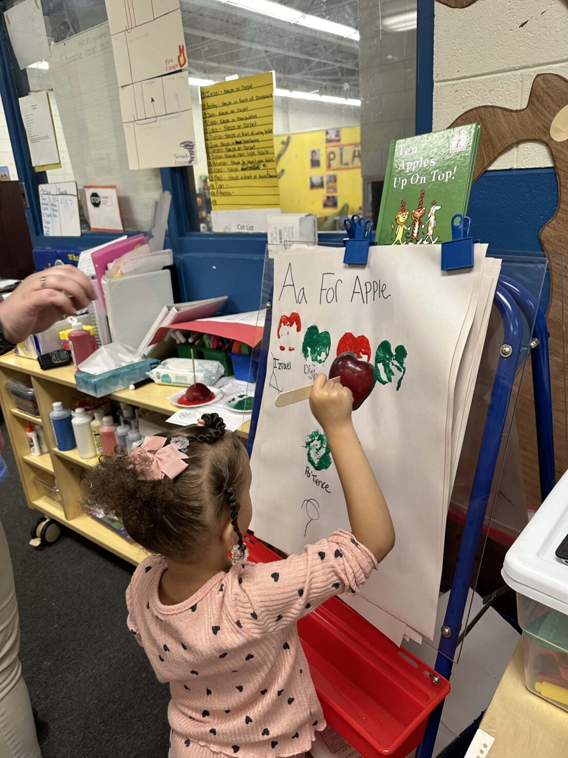 A child stands at the art easel with a stamp made out of an apple pointed at the paper. On the paper are red and green stamp imprints from the apple.