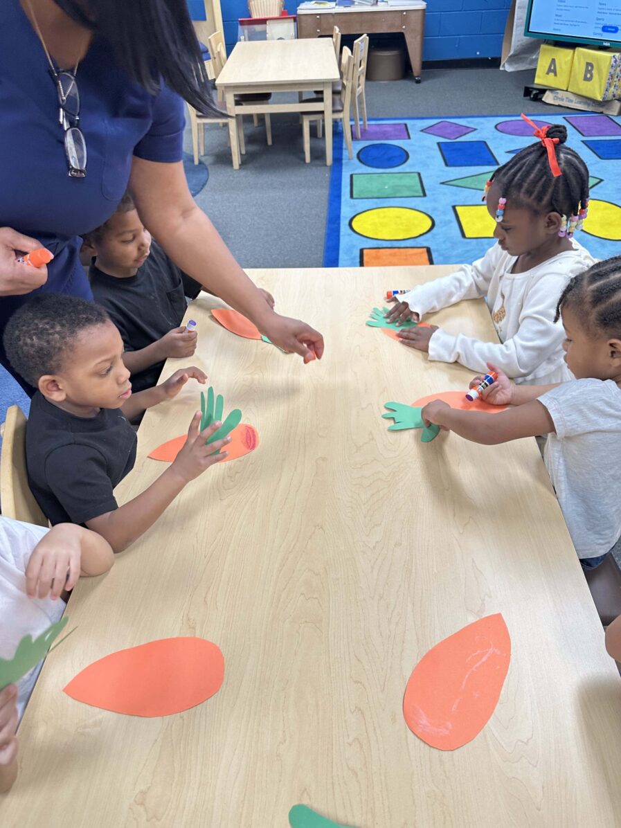 Students are sitting at their desk making cut out pictures of a carrot. The carrot top is the color green and shaped like the students hands. The body of the carrot is the color orange and shaped like a teardrop.