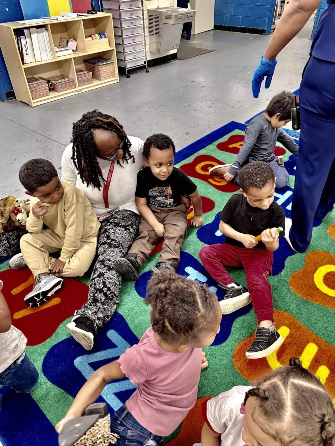 Child is holding and looking at the carrot while other students and a teacher are watching