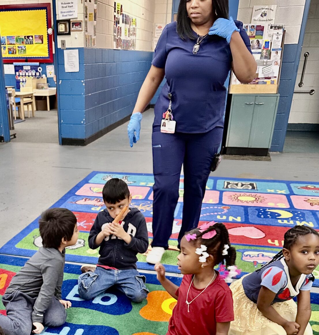 Child is smelling the carrot while other students are sitting on the rug with him.