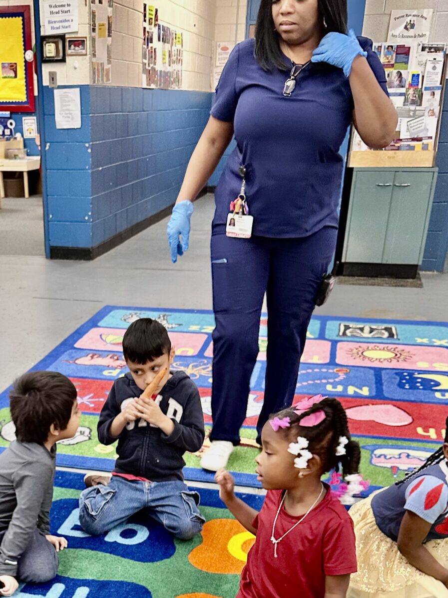 Child is smelling the carrot while other students are sitting on the rug with him.