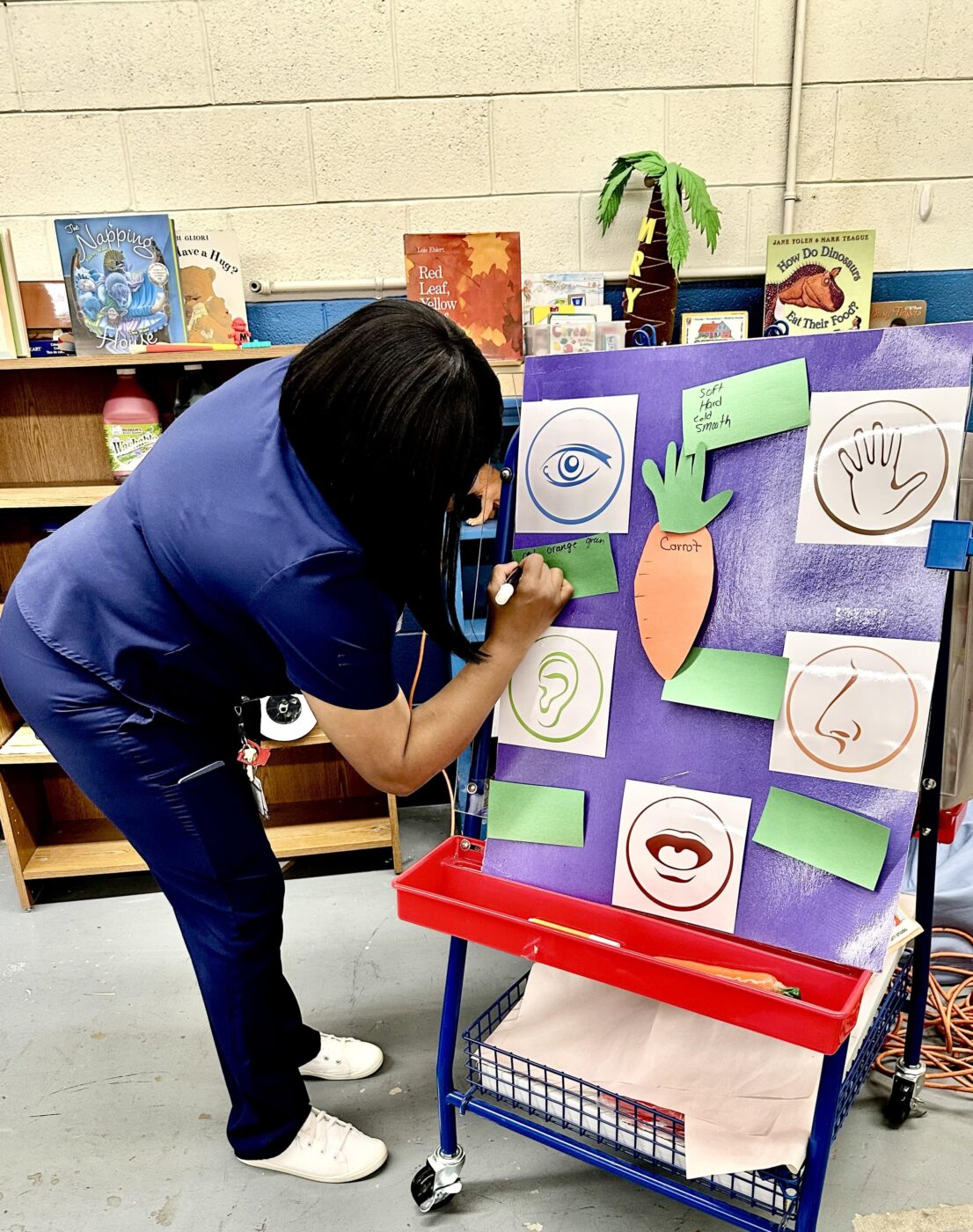 Teacher is writing down what the students are telling her about how the carrot looks, how it smells, how it tastes, and how it feels