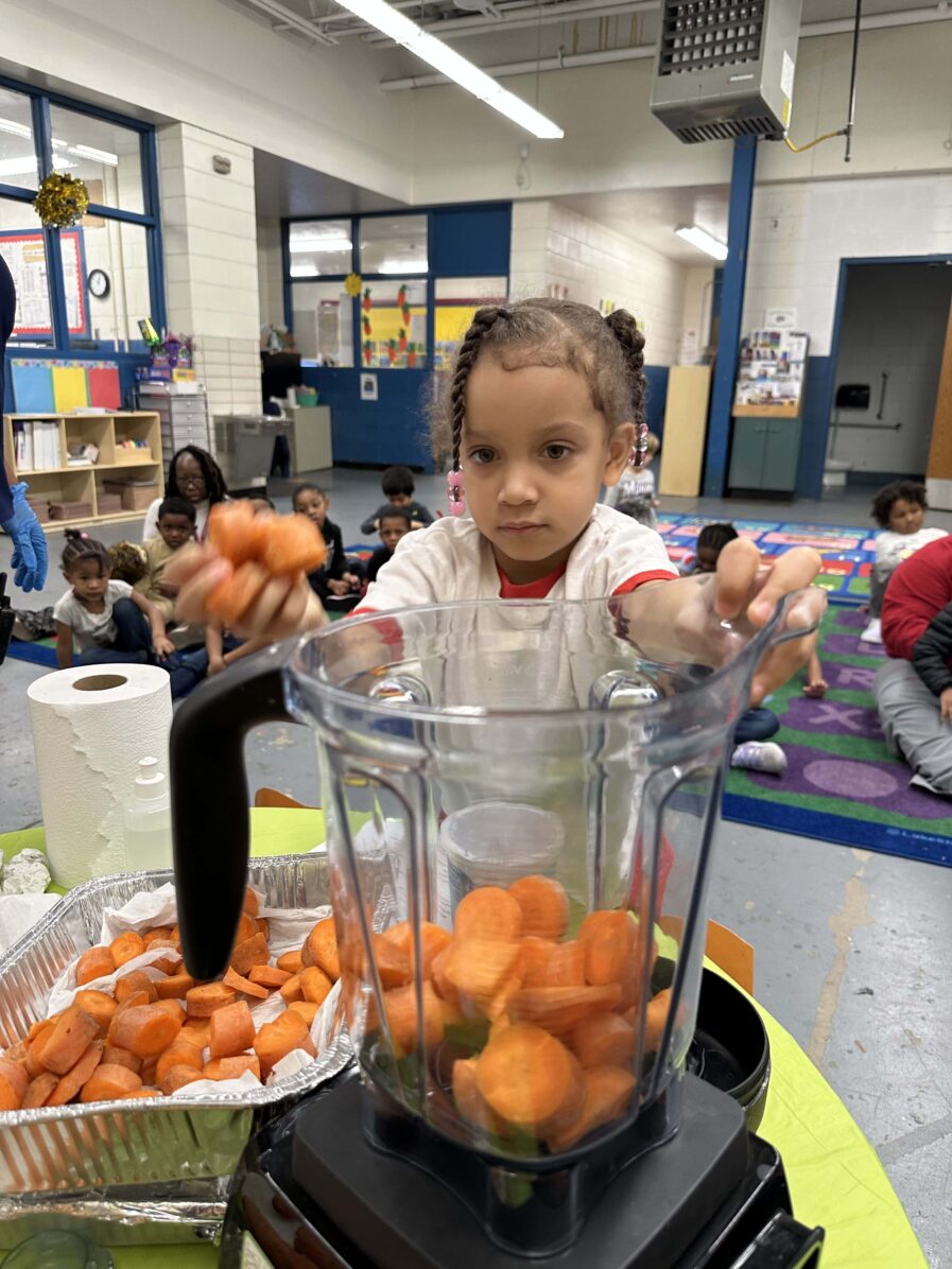 Child is putting cut pieces of carrot into a blender to make a smoothie