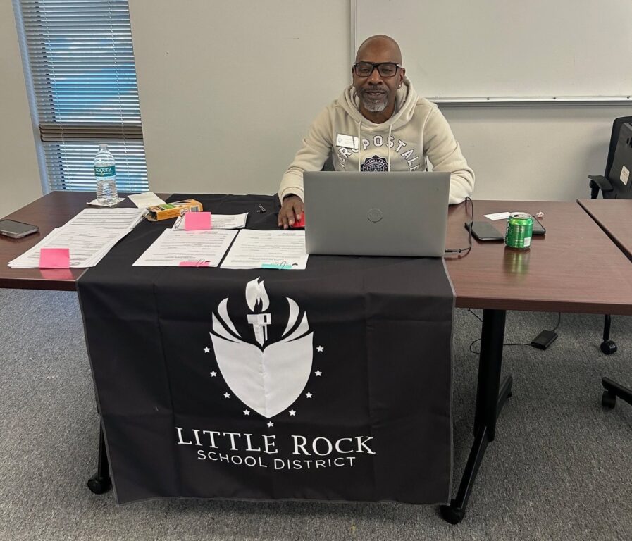 A gentleman from Little Rock School District is sitting a table with his computer while waiting to help families with enrollment.