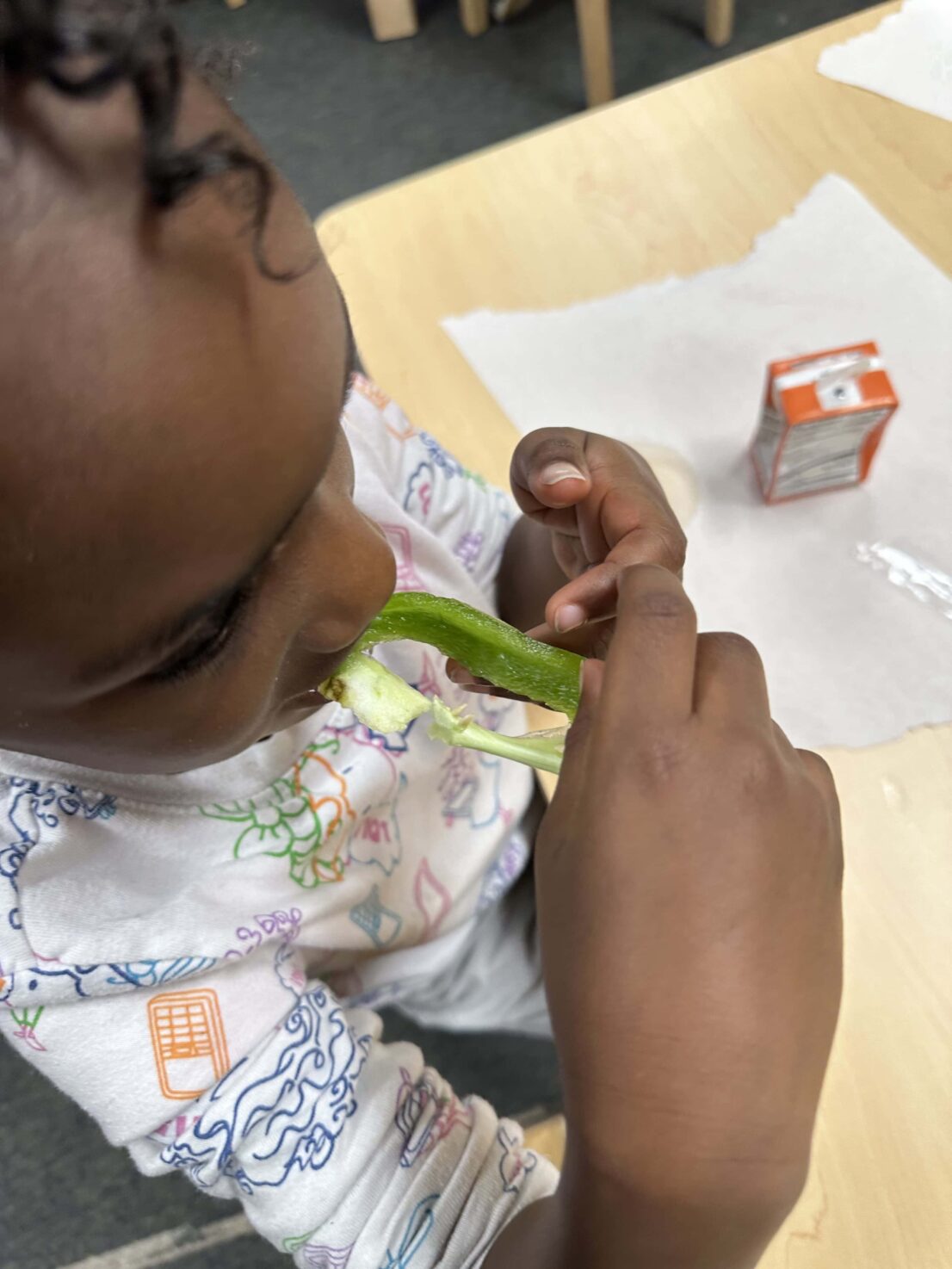 A student is tasting a slice of the green bell pepper while sitting at the desk.