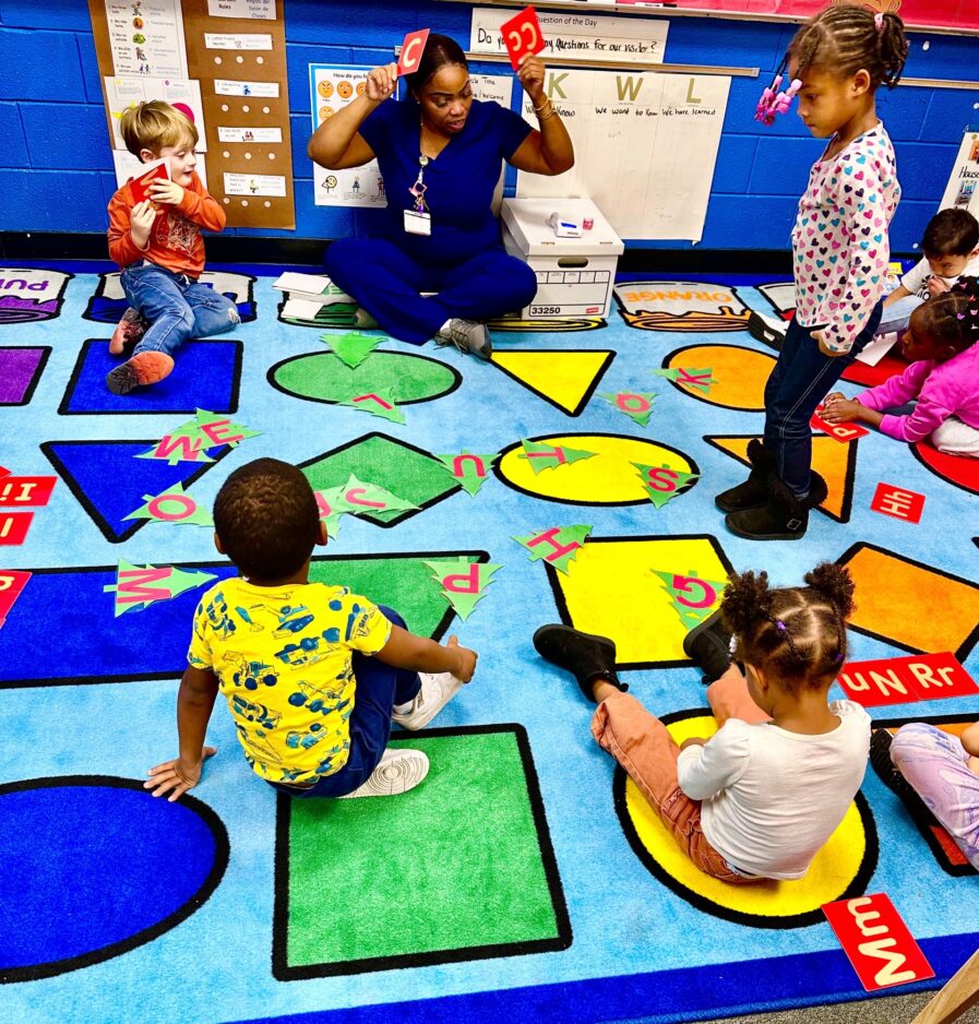 Ms. Williams is sitting on a floor rug with several children. The rug is a light blue with different colored shapes. There are circles, triangles, squares, and diamond shapes on the rug.